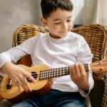 Child joyfully playing a ukulele while sitting inside, showcasing musical expression.
